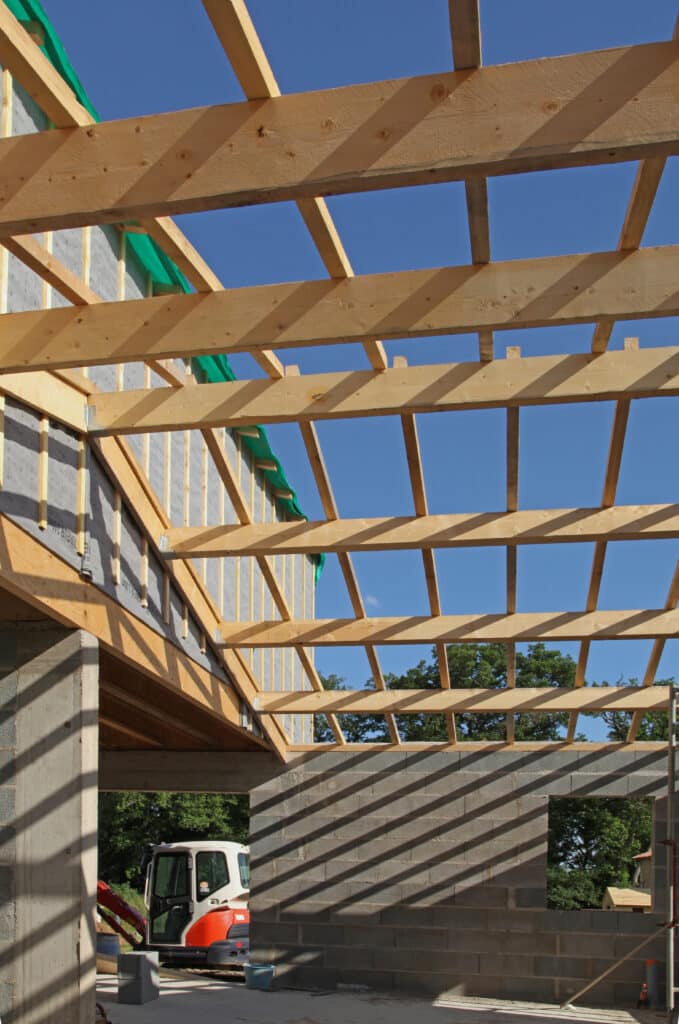 Construction Maison : Charpente Bois et Gros Œuvre Charpente bois neuve d'une construction, vue par le bas, sur fond de ciel bleu et blocs de béton. Pelleteuse visible.