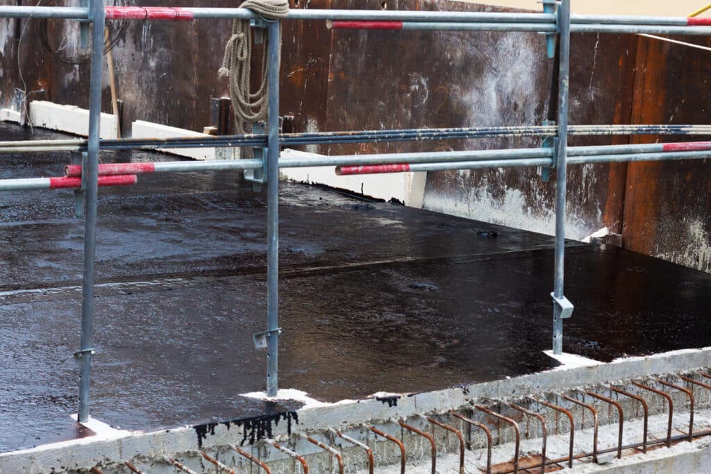Travaux de construction : étanchéité et sécurité Chantier : garde-corps de sécurité tubulaire sur une surface noire humide, avec armature en acier rouillé visible.