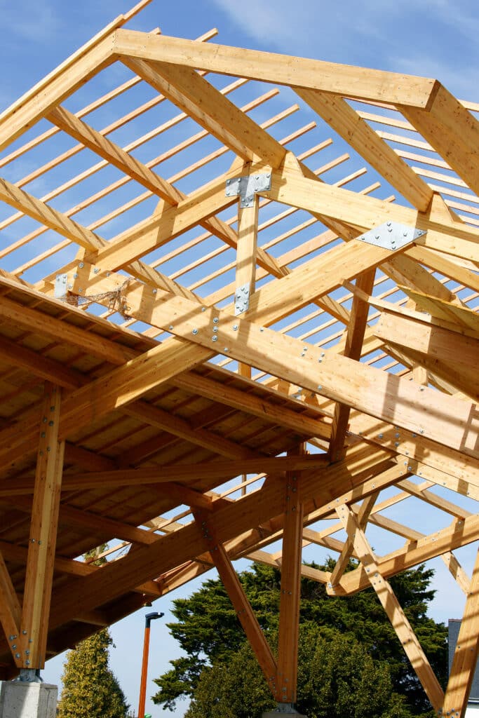 Charpente Bois Lamellé-Collé : Construction Moderne Ossature de toit en bois lamellé-collé avec assemblages métalliques apparents, vue par en dessous sous un ciel bleu.