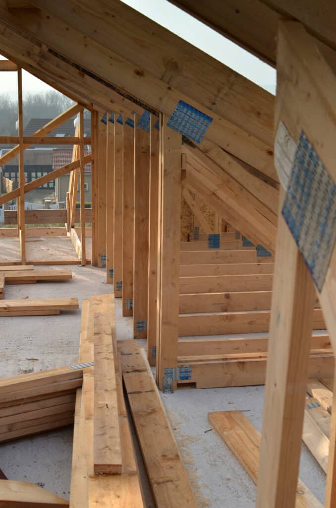 Construction Charpente Bois Maison Ossature Vue Intérieure Charpente en bois d'une maison en construction, vue de l'intérieur, avec planches au sol et maisons voisines visibles à travers l'ouverture.