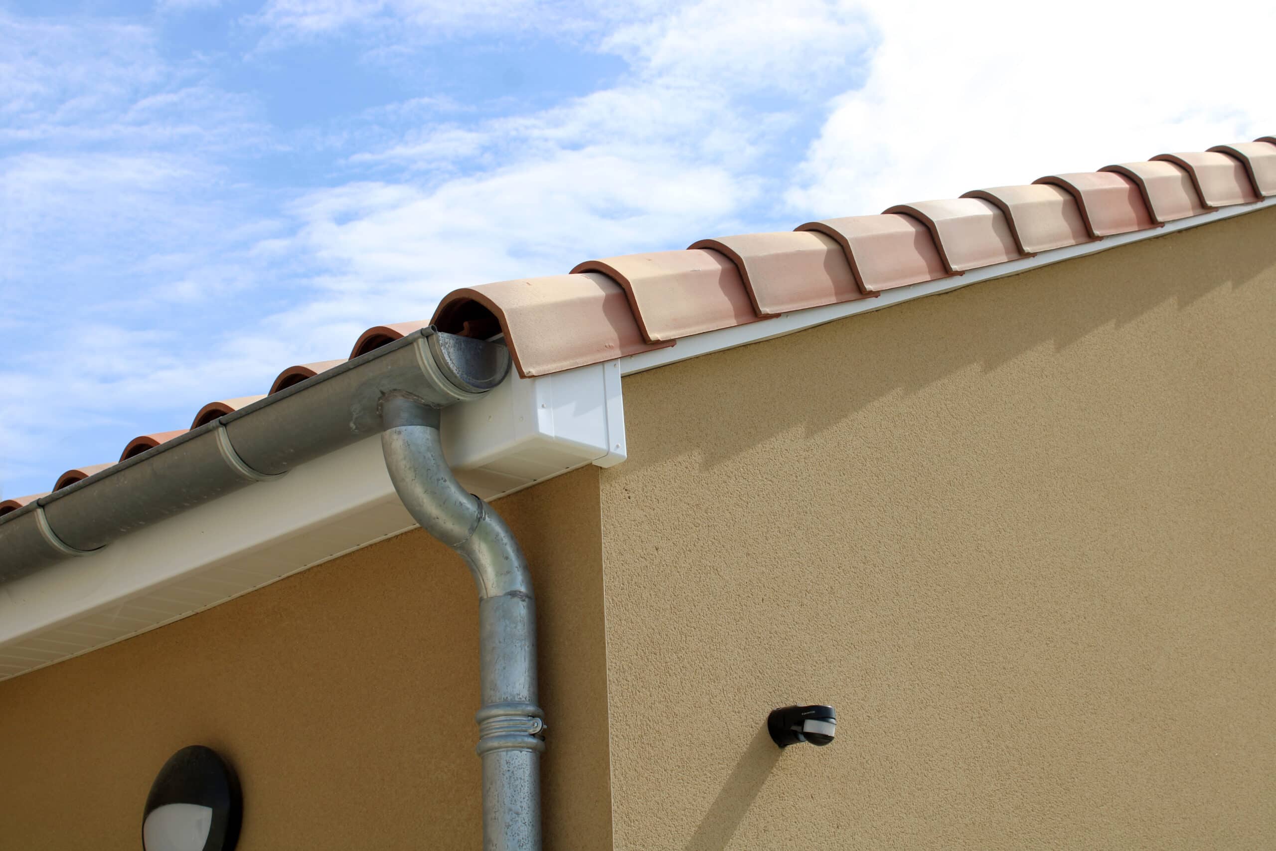 Détail d'un pignon de maison neuve avec gouttière en zinc, sous-face blanche, tuiles canal rouges et crépi ocre, sous ciel bleu nuageux. Un détecteur de mouvement est visible.