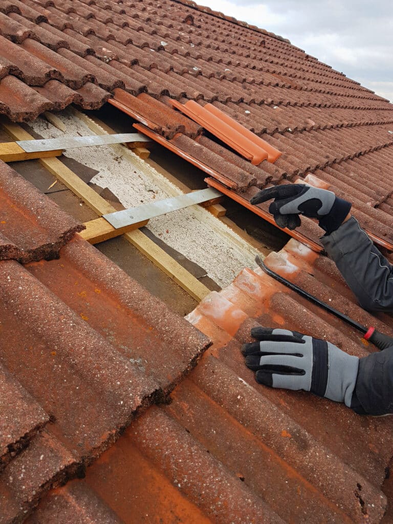 Réparation toiture tuiles : retrait d'une tuile Rénovation de toiture : ouvrier avec gants gris enlevant une tuile en terre cuite rouge pour accéder aux liteaux de bois et à l'isolant.